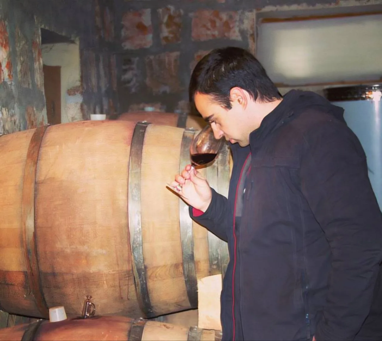 Hayk Mkrtchyan nosing a glass of red wine beside oak barrels in the Aparteny cellar